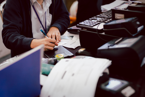 A person sitting at a desk, writing on papers with a pen. The desk is cluttered with documents, a calculator, and office equipment. The person is wearing a lanyard and a dark jacket.