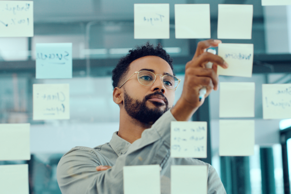 A man standing in front of a glass wall covered with sticky notes. He is wearing glasses and writing on one of the notes with a marker, appearing focused on planning or brainstorming.